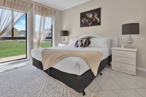 Een slaapkamer met een groot bed, een nachtkastje, een dressoir, een raam met gordijnen en een schilderij aan de muur.