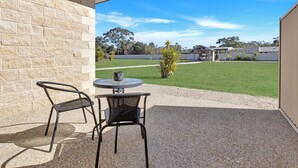 Un patio con dos sillas y una mesa, un muro de ladrillo, y vistas a un campo con césped y árboles.