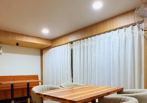 A room with a wooden dining table, cushioned chairs, a sofa, and a large window with curtains.