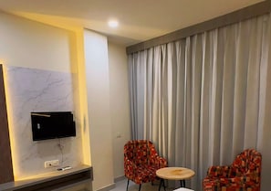 A modern room with a flat-screen TV mounted on a marble wall, two patterned chairs around a small round table, and a large window with vertical blinds.