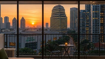 A balcony with a city view at sunset, featuring a sofa, two chairs, and a small table with a candle.