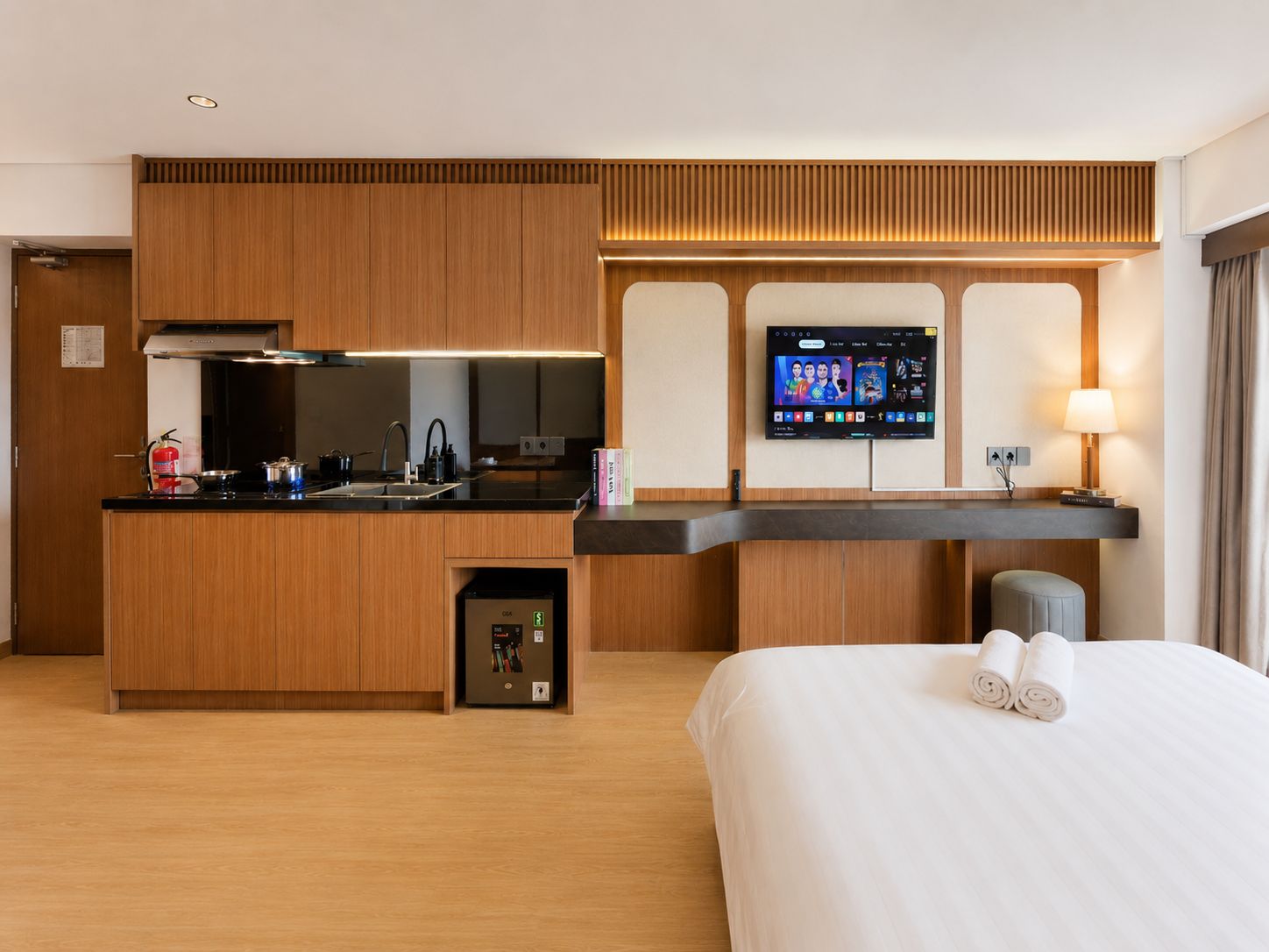Une chambre d’hôtel équipée d’une kitchenette, d’un téléviseur à écran plat, d’une table de chevet avec une lampe et d’un lit avec des serviettes.