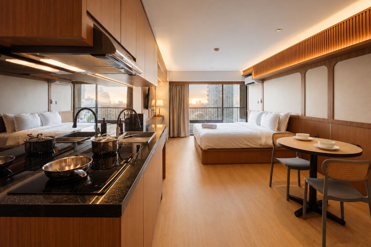 A modern hotel room with a kitchenette, a bed, a dining area, and a large window with a city view.