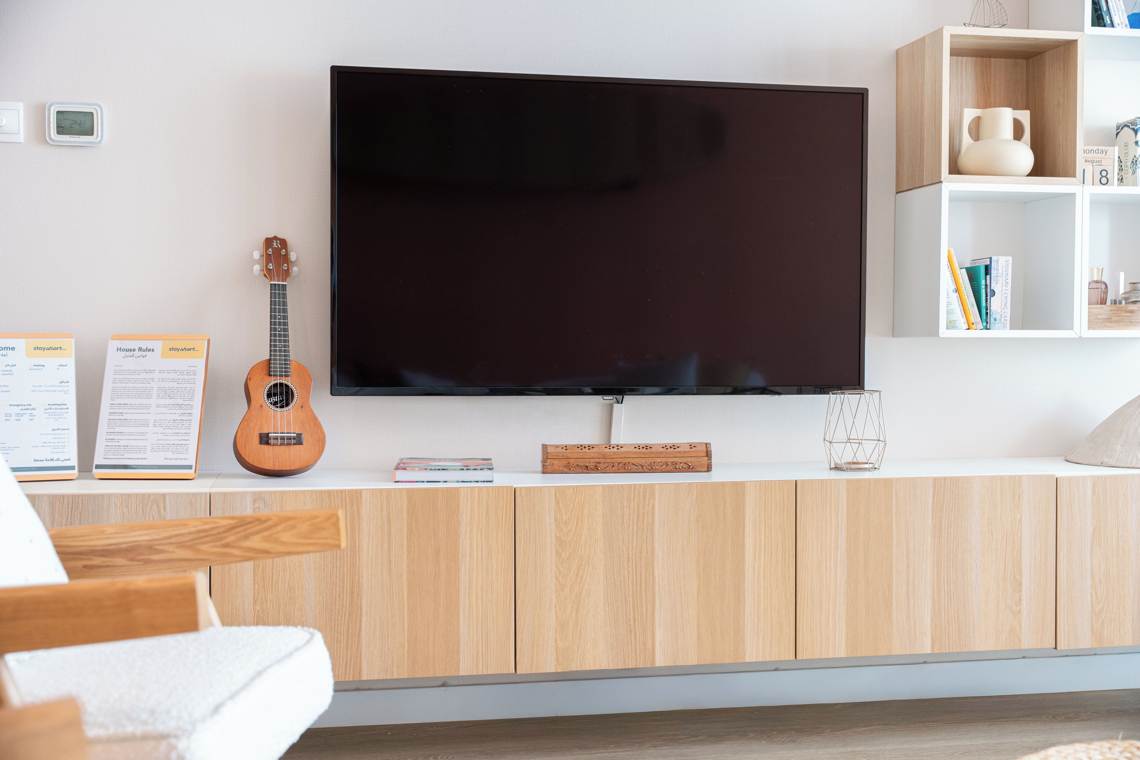 A modern living room with a flat-screen TV mounted on a wooden cabinet, a ukulele, and a bookshelf with books and decorative items.