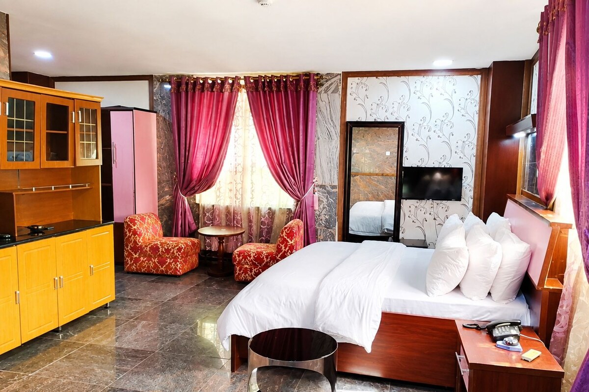 A hotel room with a bed, a desk, a chair, a small table, a cabinet, and a television.
