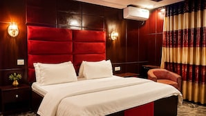 A hotel room with a large bed, two bedside tables, a nightstand, a red upholstered headboard, wall sconces, an air conditioning unit, and a patterned curtain. - Galaxy La Palm Royal Resort Abakaliki  (Abakaliki)