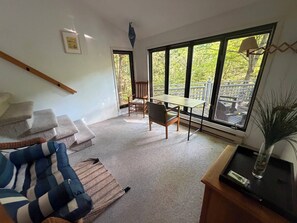 A room with a sofa, a table, and chairs, large windows with a view of trees, and a wooden ceiling beam.
