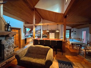 A spacious living area with a stone fireplace, wooden ceiling beams, a dining table with chairs, and a kitchen in the background.