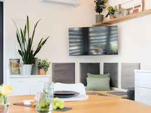 A modern living room with a dining table, a sofa, a TV mounted on the wall, and various decorative plants.
