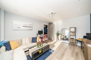Ein modernes Wohnzimmer mit Sofa, Couchtisch und Fernseher. Außerdem ein Bücherregal und ein Schreibtisch mit Computer.