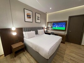 Quarto de hotel moderno com cama grande, mesa de cabeceira e TV na parede.