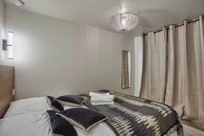 Een keurig opgemaakt bed met witte lakens en twee donkere kussens, een bedlampje, een spiegel en een raam met beige gordijnen.