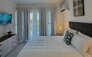 Un dormitorio con cama, televisor, cómoda, mesita de noche con lámpara y ventana con cortinas.