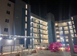 A multi-story building with balconies, a covered outdoor area, and a landscaped garden with pink flowers. - 1BR Comfort City Apartment with pool (Kingston)