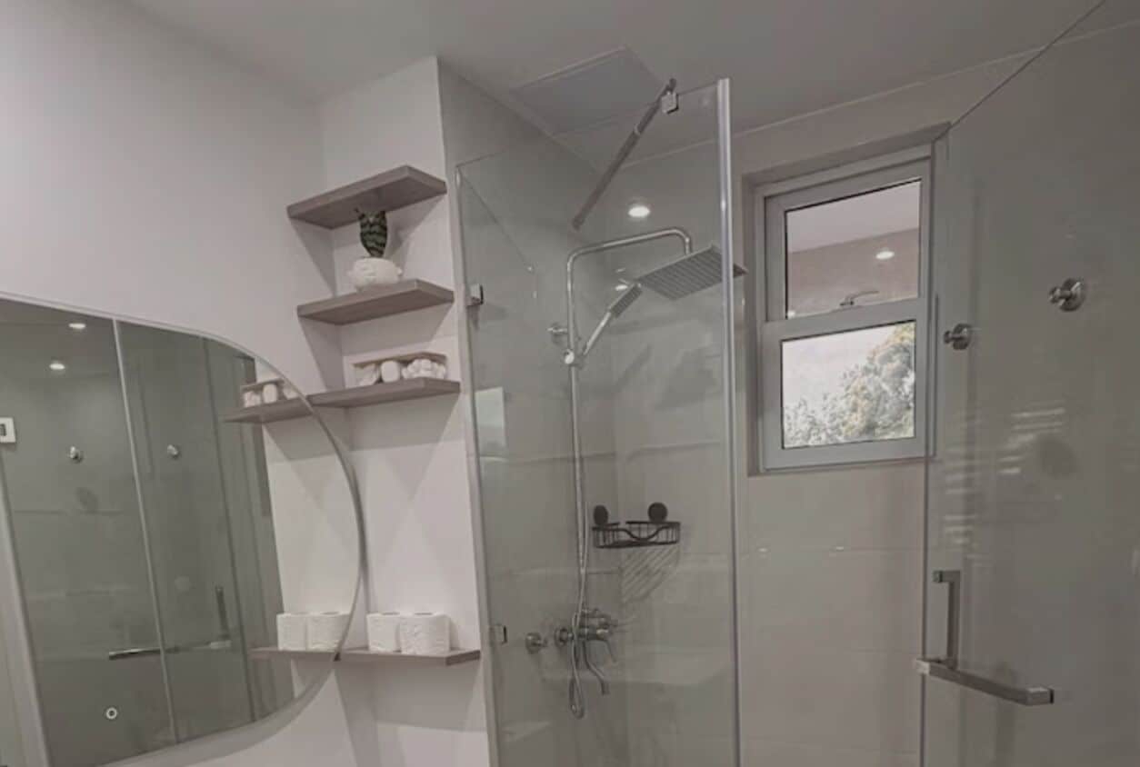A modern bathroom with a glass-enclosed shower, a toilet, and a sink. There are shelves with toiletries and a window above the shower.