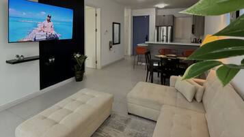 Una sala de estar con televisor de pantalla plana, un sofá, un comedor con sillas y una cocina al fondo.