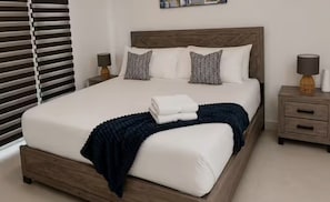 A neatly made bed with a dark blue throw blanket and pillows, a wooden headboard, and bedside tables with lamps. - 1BR Comfort City Apartment with pool (Kingston)