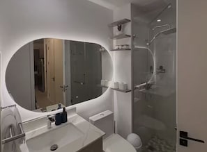 A modern bathroom with a large oval mirror, a glass-enclosed shower, and a toilet. - 1BR Comfort City Apartment with pool (Kingston)