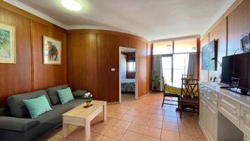 Una sala de estar con sofá, mesa de centro y televisor. Incluye un área de cocina con armarios y una mesa de comedor.