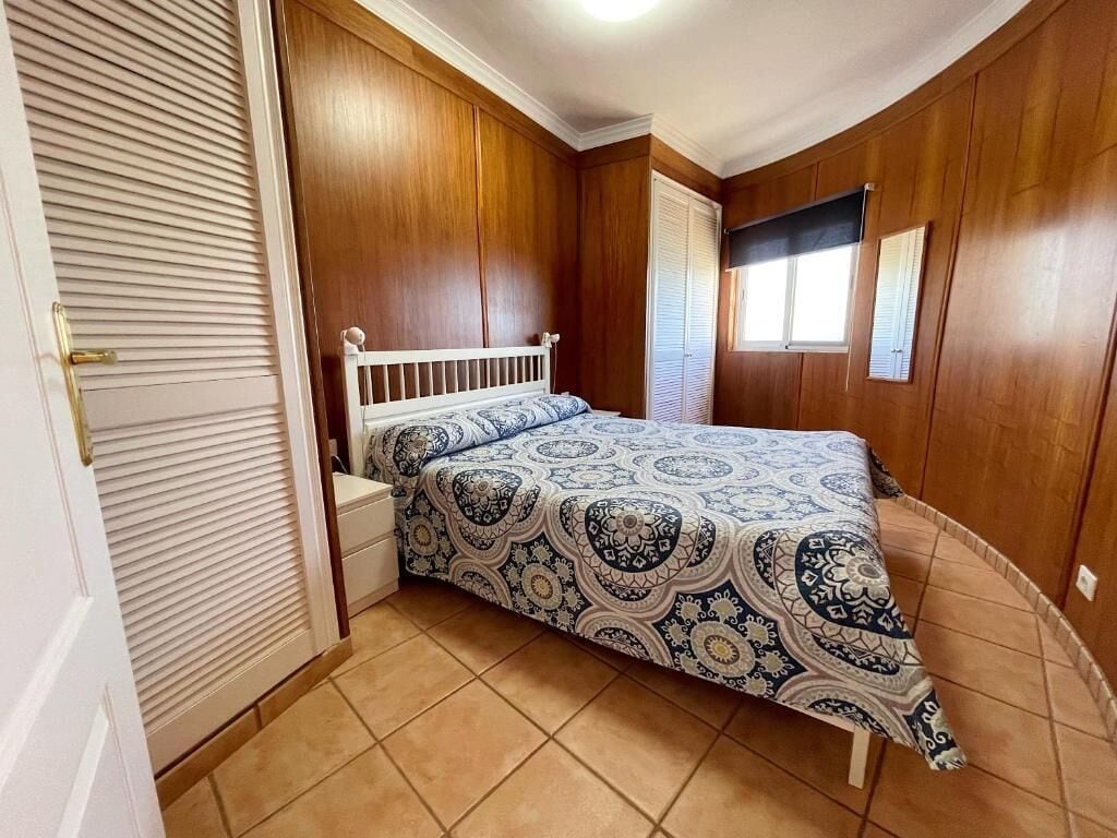 Un dormitorio con cama, mesita de noche, ventana con persianas y paredes de madera.