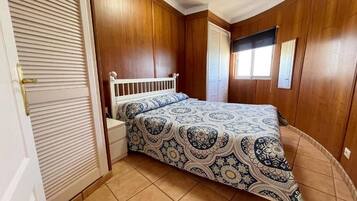 Un dormitorio con cama, mesita de noche, ventana con persianas y paredes de madera.