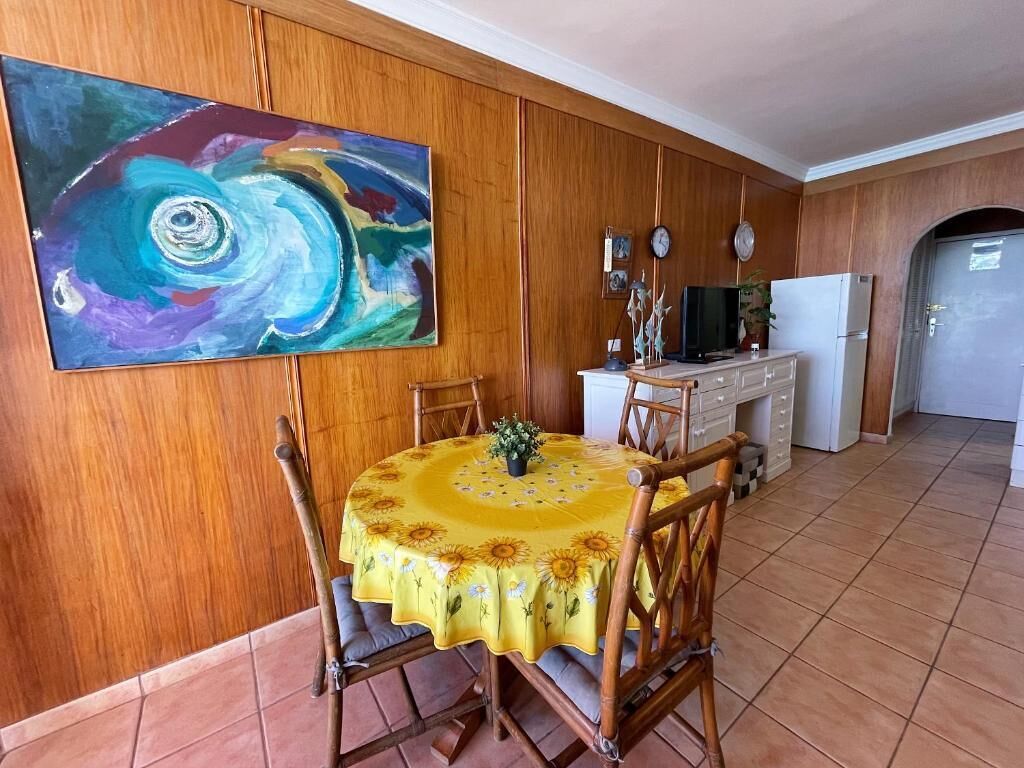 A dining area with a round table covered by a yellow tablecloth, wooden chairs, and a painting on the wall.