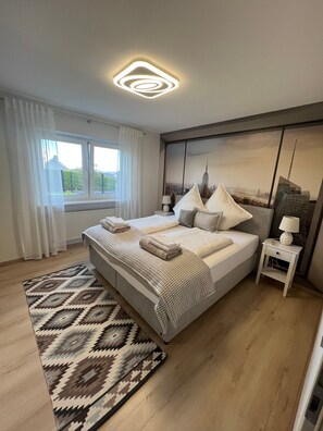 A modern bedroom with a large bed, a nightstand, a window with curtains, and a cityscape view.