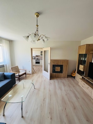 Ein modernes Wohnzimmer mit einem Glastisch, einer Couch, einer Holzkommode und einer durchgehenden Tür sichtbaren Küche.