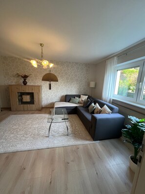 A modern living room with a sectional sofa, a glass coffee table, a wooden cabinet, a fan, and a large window.