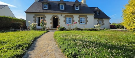 Une maison à deux étages, avec un chemin de pierre menant à l’entrée, entourée d’un jardin bien entretenu où poussent différentes sortes de fleurs.