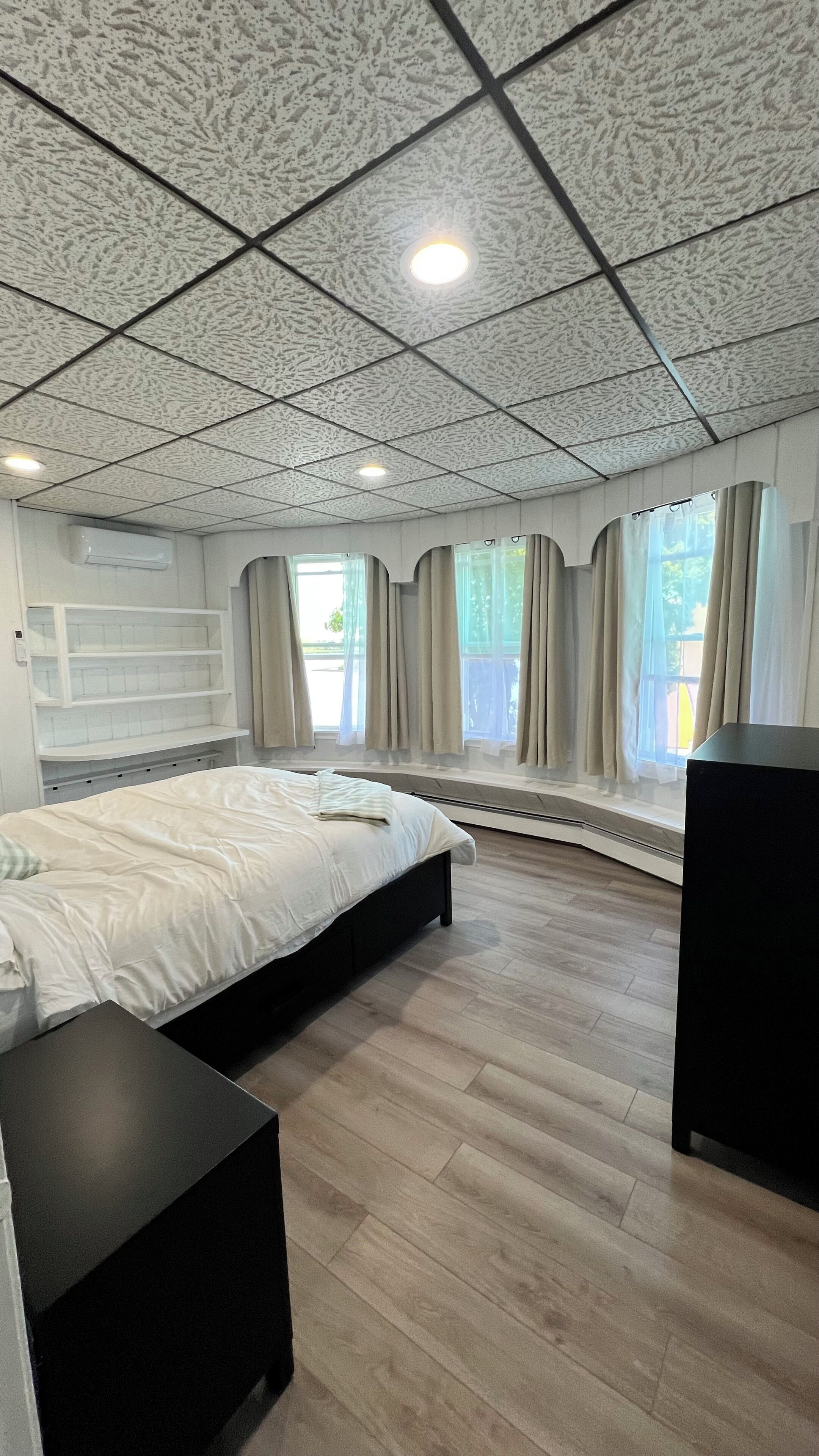 A bedroom with a bed, a black nightstand, a built-in bench, a window with curtains, and a ceiling with a grid pattern.