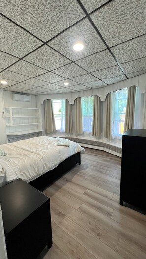 Ein Schlafzimmer mit einem Bett, einem schwarzen Nachttisch, einer eingebauten Bank, einem Fenster mit Vorhängen und einer Decke mit Gittermuster.