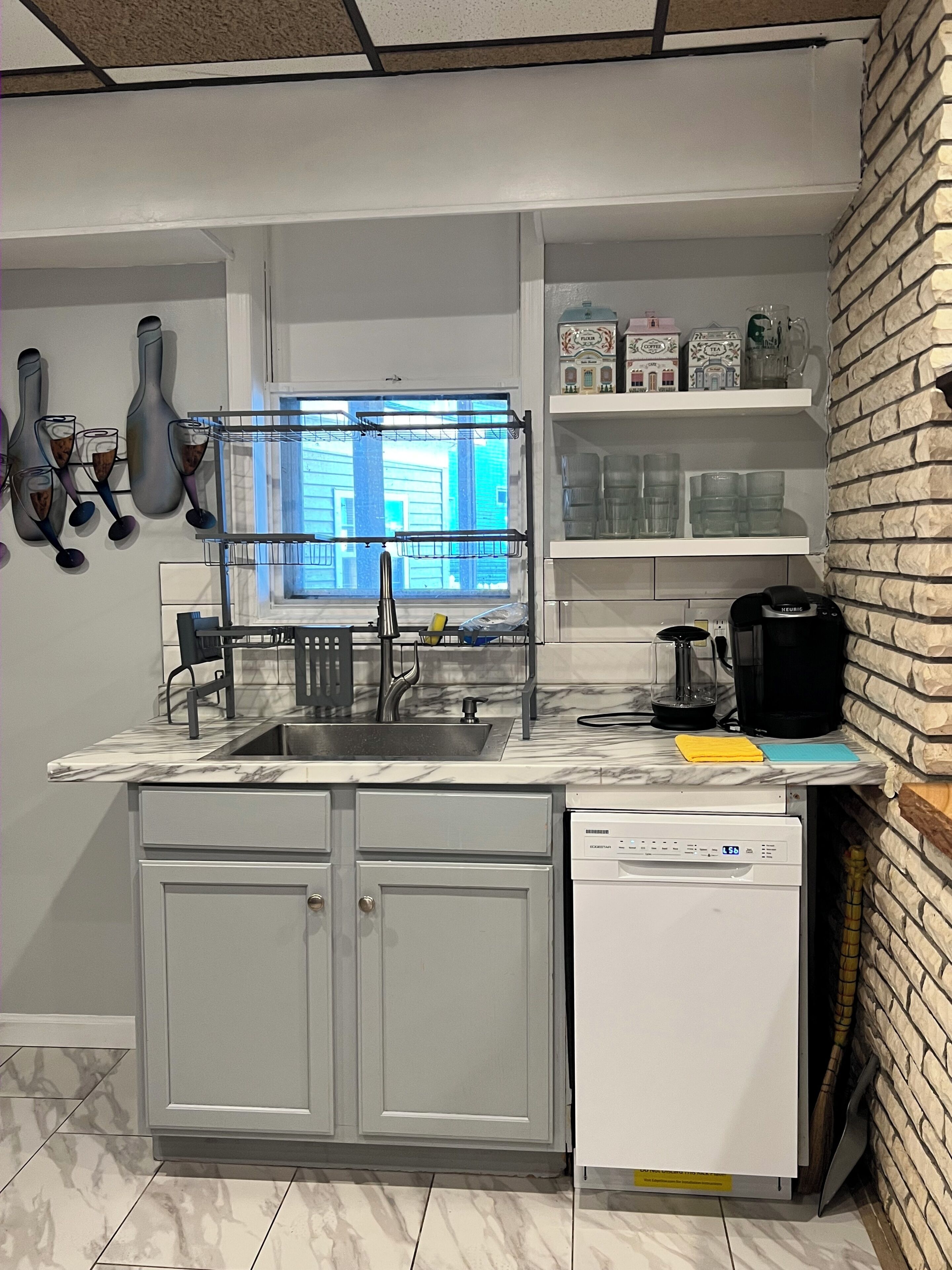 A compact kitchen with a built-in dishwasher, a window with blinds, and a brick accent wall.