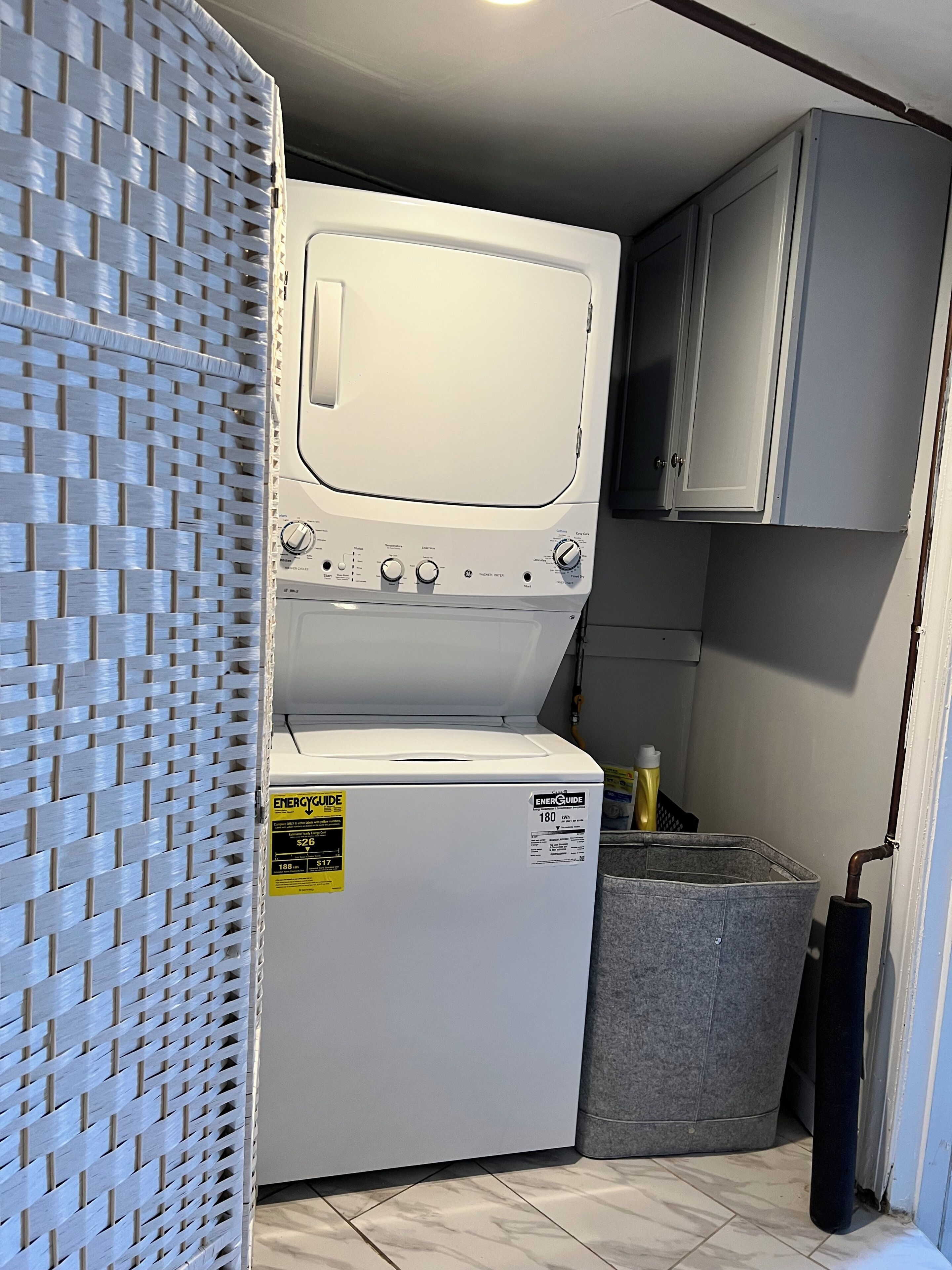 Een kleine wasruimte met een voorlader wasmachine en droger, een grijze opbergkist en een wandstructuur van gevlochten materiaal.