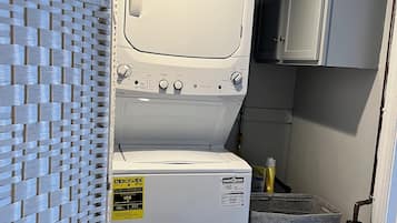 A compact laundry area with a front-loading washer and dryer, a grey storage bin, and a woven partition.
