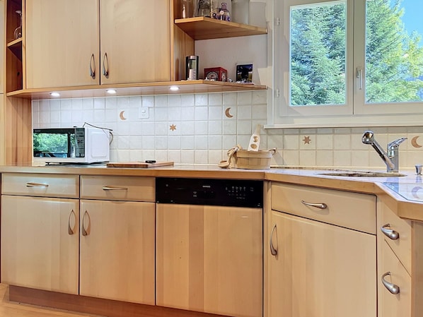 Een keuken met houten kastjes, een magnetron, een vaatwasser en een raam met uitzicht op bomen.