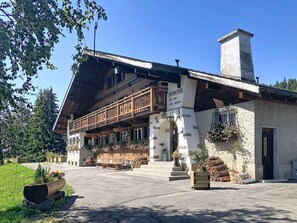A traditional chalet with a wooden balcony, a stone chimney, and a sign reading 'Anbärg des Jous'.