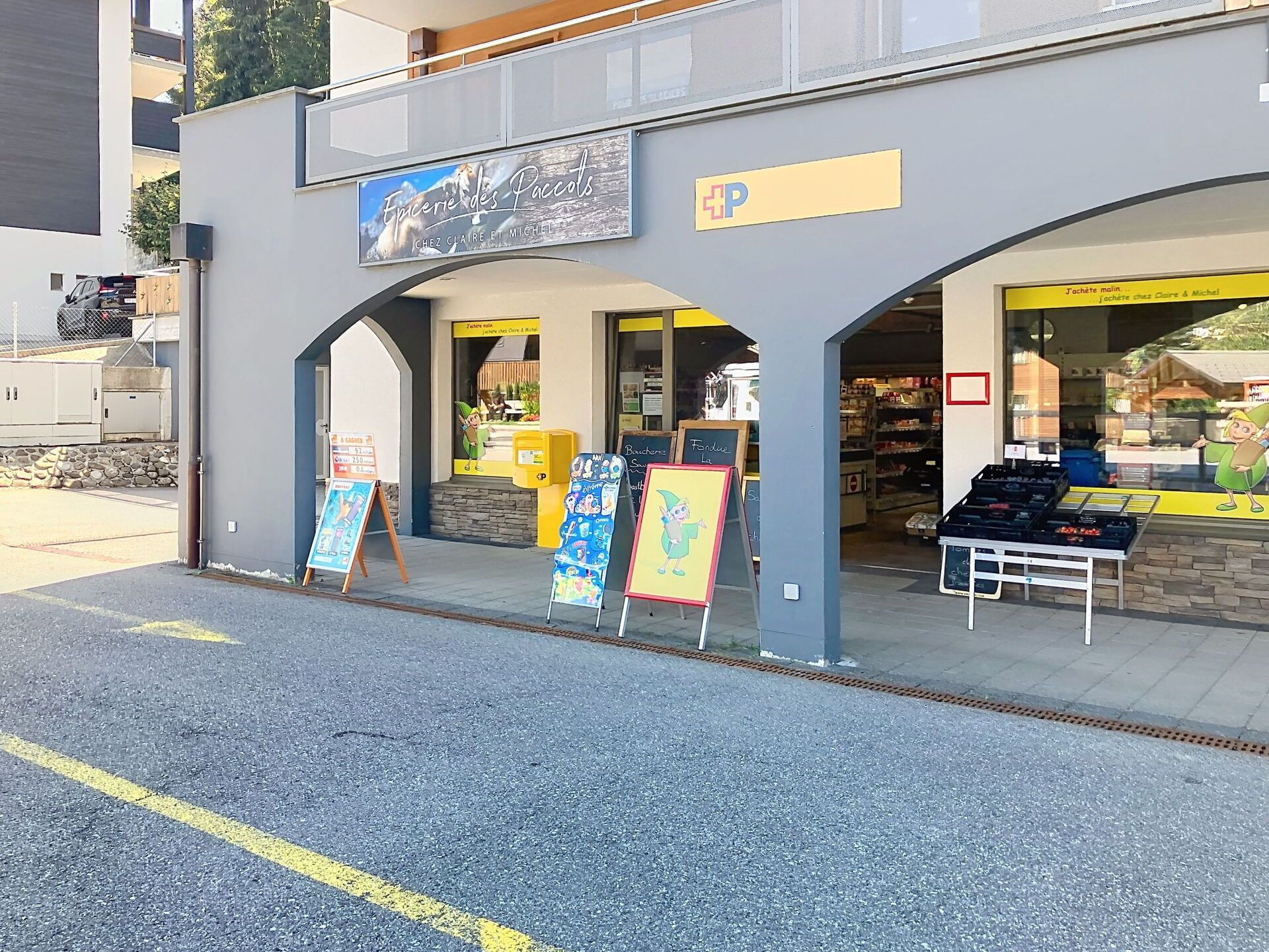 Fachada de loja com uma placa indicando 'Epicerie des Paccol's' e uma placa amarela com um símbolo de mais. Há expositores externos e uma entrada visível.