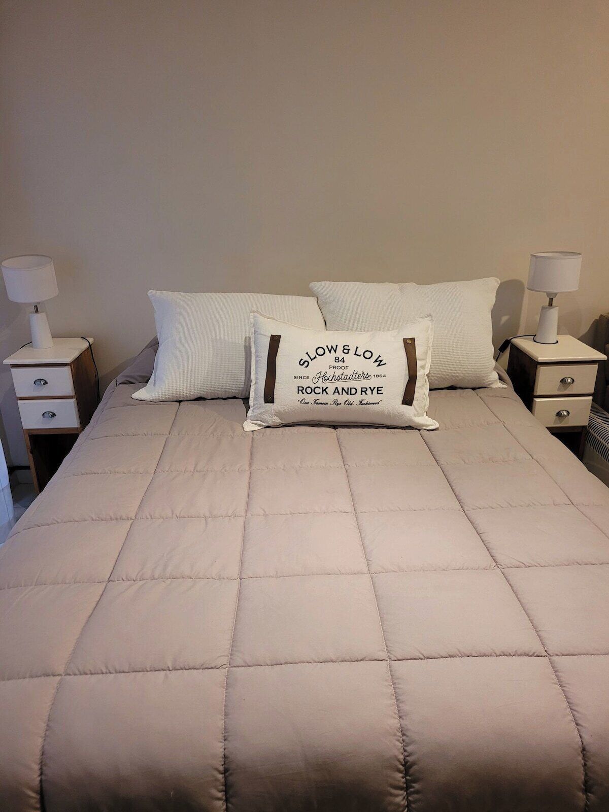 Un lit recouvert d’une courtepointe beige matelassée, deux oreillers blancs et deux tables de chevet avec des lampes.