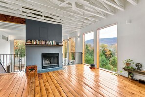 A spacious room with a fireplace, wooden flooring, large windows, and a view of a mountainous landscape. - The Windbourne A Luxury Retreat In The Catskills (Saugerties)