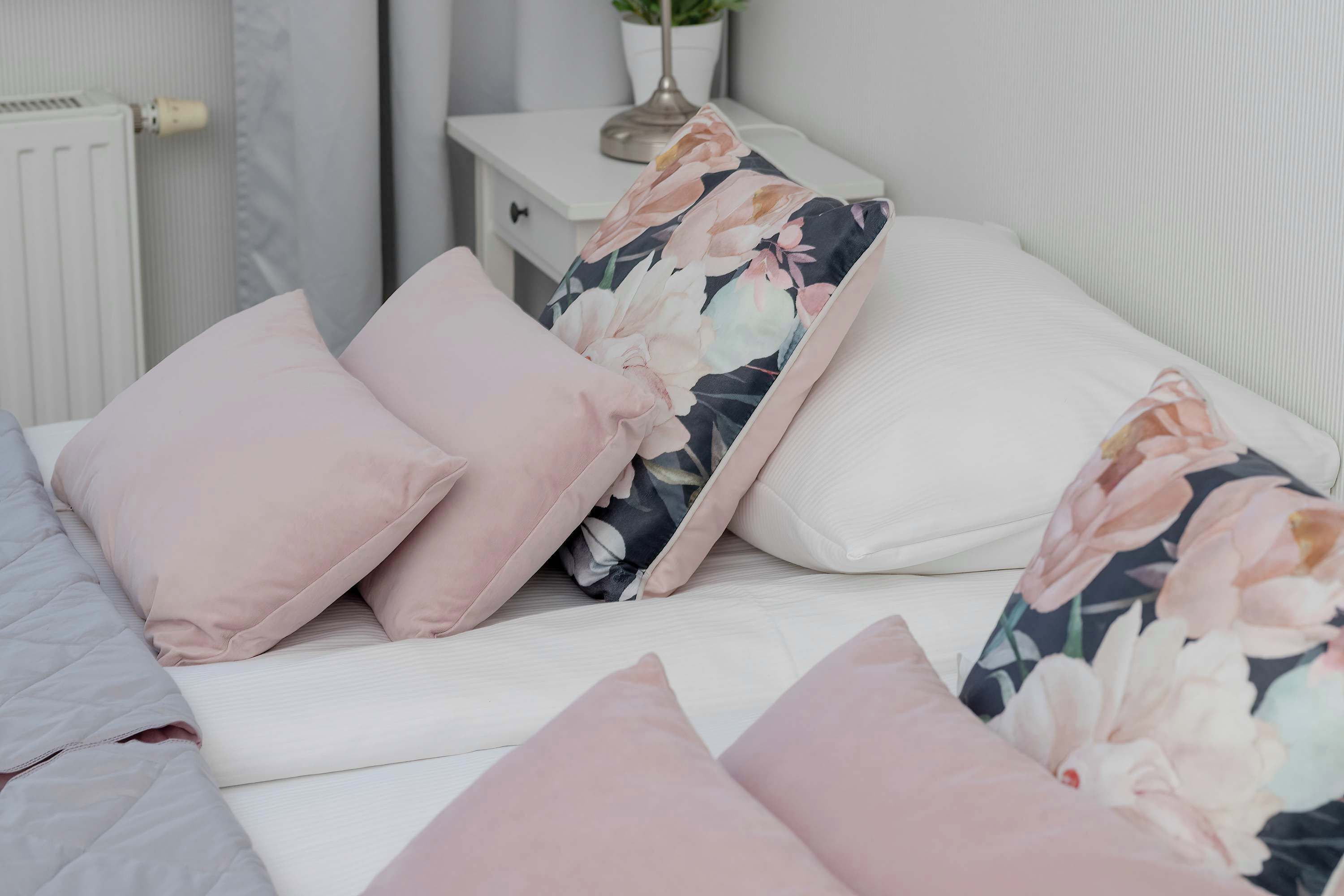 Una cama bien hecha con almohadillas rosas y estampadas, una mesita de noche blanca con una lámpara y un radiador al fondo.