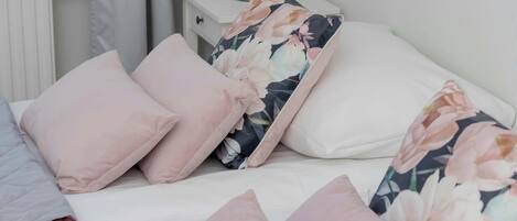 Una cama bien hecha con almohadillas rosas y estampadas, una mesita de noche blanca con una lámpara y un radiador al fondo.