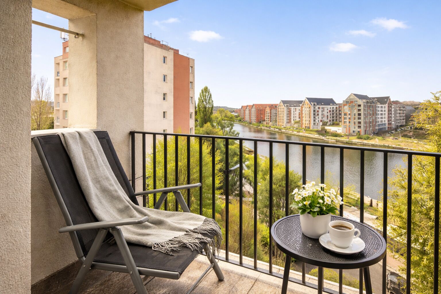 A balcony with a chair, a small table with a cup and flowers, and a view of buildings and a river.