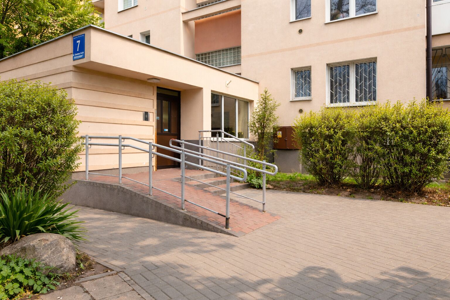 A building entrance with a ramp and metal handrails, surrounded by greenery and a paved walkway.
