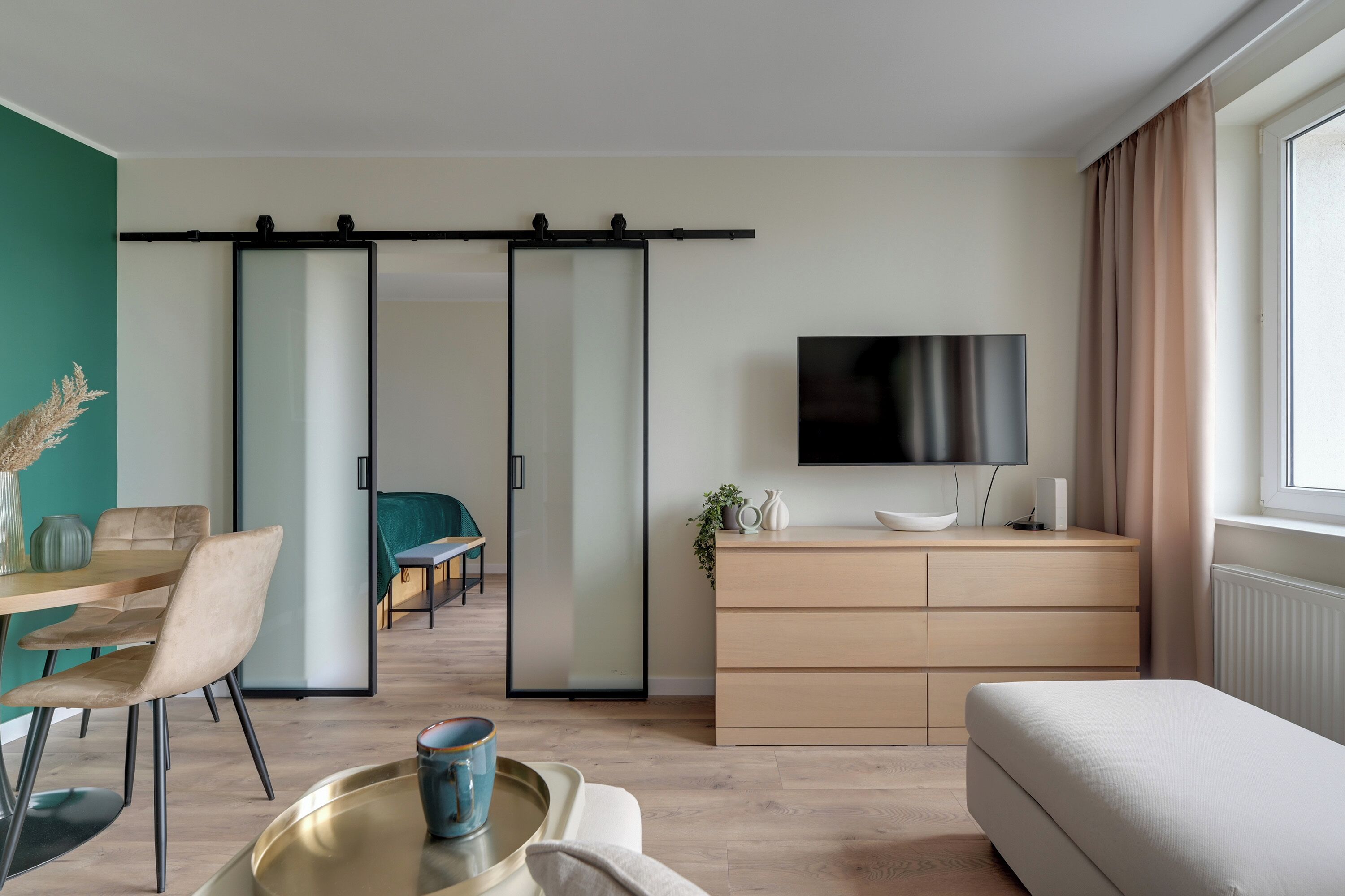 A modern living room with a sofa, a dining table with chairs, a sliding door, a TV, and a window with curtains.