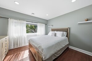 A bedroom with a bed, a dresser, a window with curtains, and a shelf with a decorative item. - Lakefront Retreat | Game Room • Pizza Oven (Hewitt)