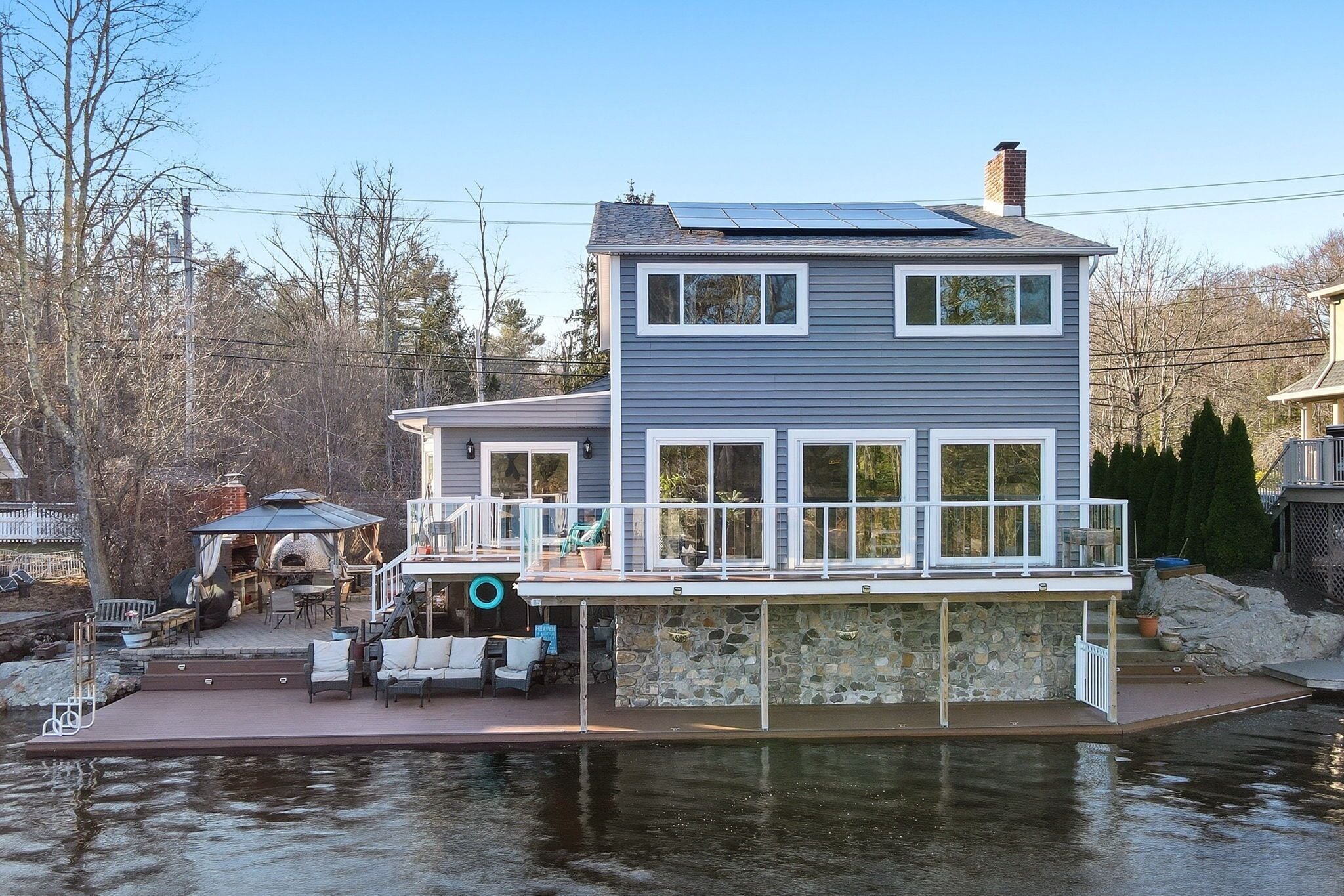 Une maison  à deux étages avec une jetée s’avançant dans un plan d’eau, un espace extérieur couvert aménagé en salon et des fondations en pierre.
