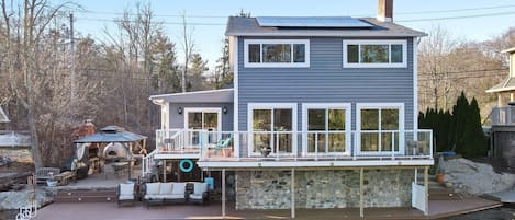 Une maison à deux étages avec une jetée s’avançant dans un plan d’eau, un espace extérieur couvert aménagé en salon et des fondations en pierre.