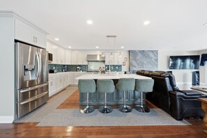 A modern kitchen with stainless steel appliances, a central island with bar stools, and a seating area with a sofa and chairs. - Lakefront Retreat | Game Room • Pizza Oven (Hewitt)