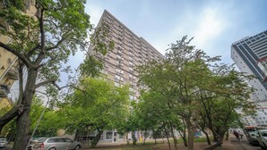 Un edificio residencial alto con varios balcones, rodeado de árboles y coches estacionados.
